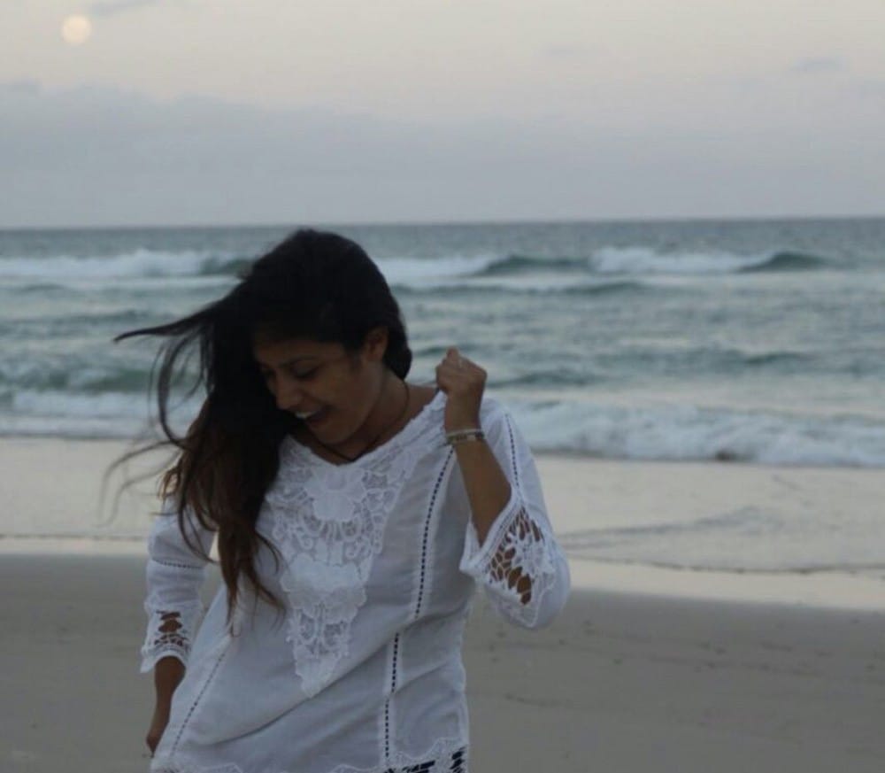 chica morena con una camisa blanca y sonriendo. Está mirando la arena y anda por la orilla del mar